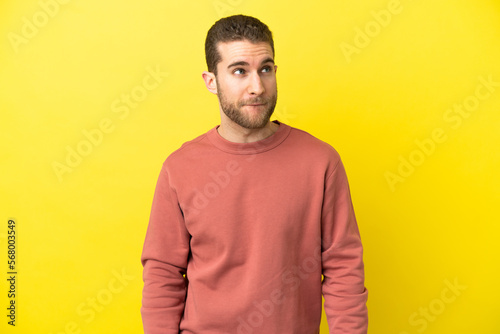 Handsome blonde man over isolated yellow background having doubts while looking up