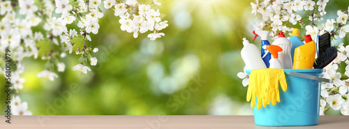 Spring cleaning. Bucket with detergents and tools on wooden surface under blossoming tree against blurred green background, space for text. Banner design