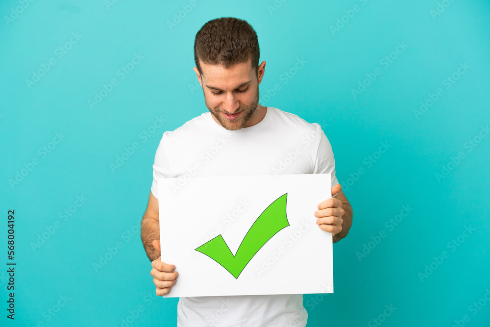 Handsome blonde man over isolated blue background holding a placard with text Green check mark icon Handsome blonde man over isolated blue background holding a placard with text Green check mark icon