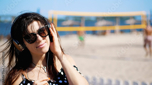 portrait, beautiful girl in sun glasses wearing big black headphones, listening to music from smartphone, on beach, on hot summer day, against background of beach volleyball