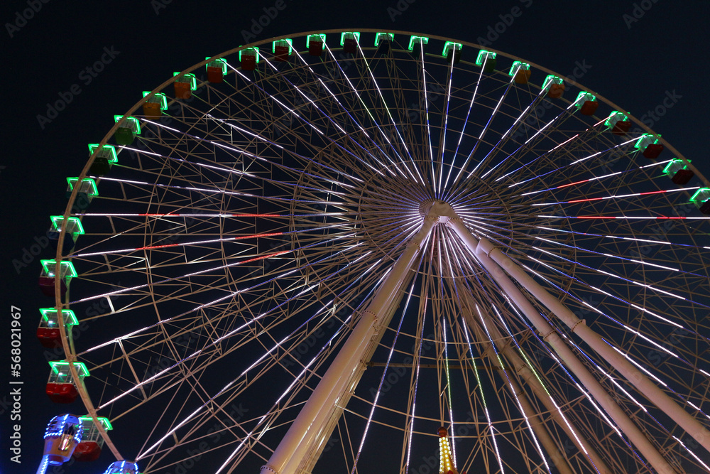 Perspective looking up at a large ferris wheel in an amusement park or funfair.