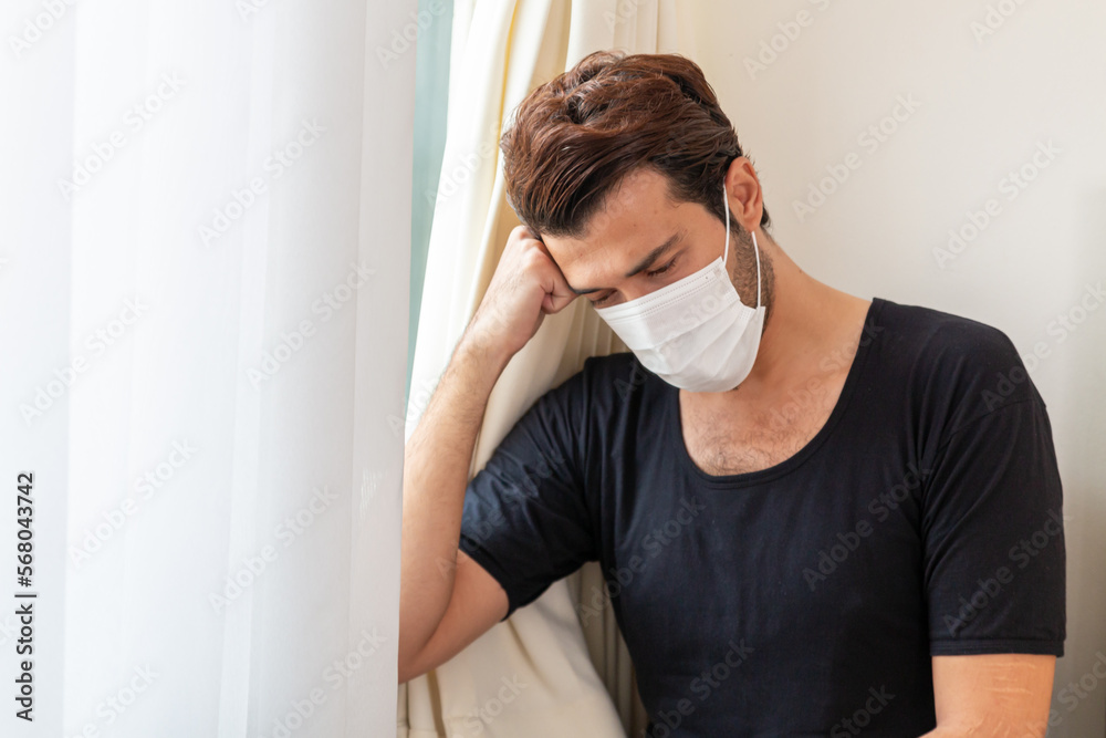 Man wearing protective mask feeling sad sitting on bed at home. Make wearing a protective mask sits on bed, with stress and headache. Man wear mask to prevent germs Covid-19. Man wearing protective mask feeling sad sitting on bed at home. Make wearing a protective mask sits on bed, with stress and headache. Man wear mask to prevent germs Covid-19.
