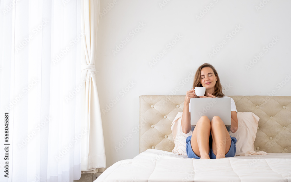 Young woman sitting on bed and using laptop. Beautiful Young woman using laptop on bed at home in the morning. Young female using laptop, sitting on bed in hotel.