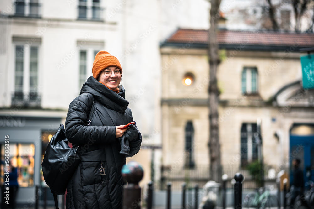 Happy smiling woman walking in Paris city in winter. Old city. Tourist. The girl is standing with the phone. The girl is wearing a black coat and an orange hat. Walking down the street.