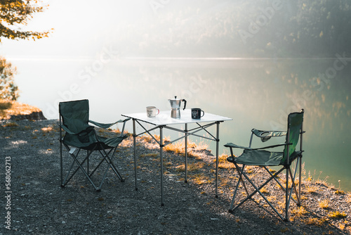Outdoor two empty chairs with picnic table and moka pot coffee for Camping against tranquil lake in the morning. Equipment for camping kitchen, cooking in nature, coffee outdoor with camping ustensils
