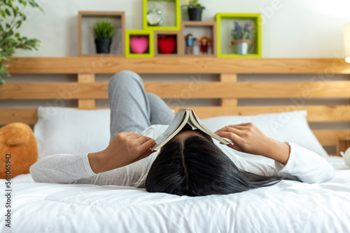 Young woman in pleasure white casual cloth lying down reading open book on bed. Asian girl imagine story and smiling about novel book.