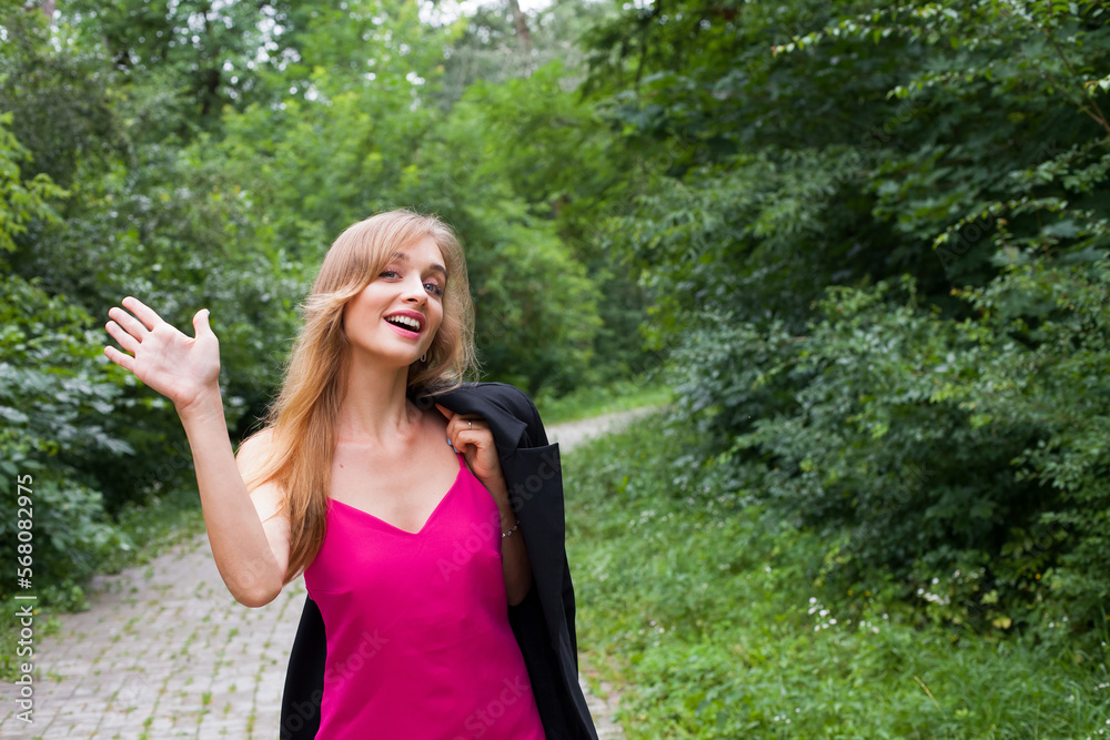 A slender young girl with long blond hair, in a short stylish pink dress, walks beautifully along the path in the forest in summer. She is holding a jacket on his shoulder. Smiling and waving. A slender young girl with long blond hair, in a short stylish pink dress, walks beautifully along the path in the forest in summer. She is holding a jacket on his shoulder. Smiling and waving.