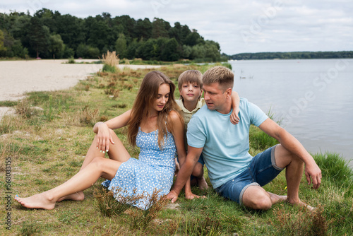 Portrait of a happy family in nature, on the banks of a picturesque river. Mom is dressed in a summer dress, dad and son are in shorts and t-shirts.