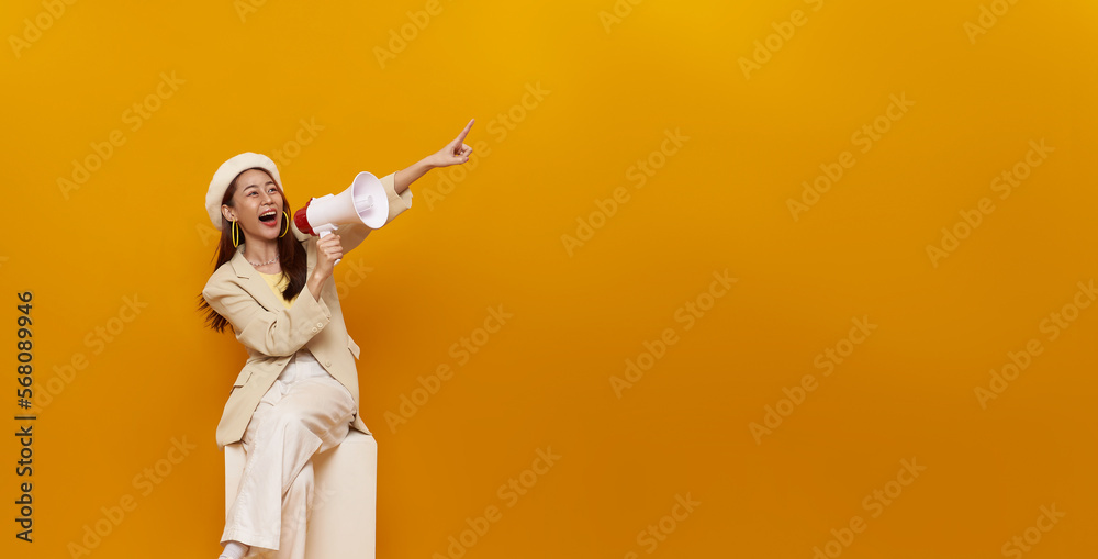 Young Asian woman sitting on white box holding megaphone and pointing finger isolated on yellow background. Speech and announce concept. Young Asian woman sitting on white box holding megaphone and pointing finger isolated on yellow background. Speech and announce concept.