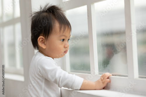 Lonely Adorable asian baby toddler standing in front of windows and looking outside waiting mom or dad come back home.Sad Baby standing looking outside home alone want to play with friends outside