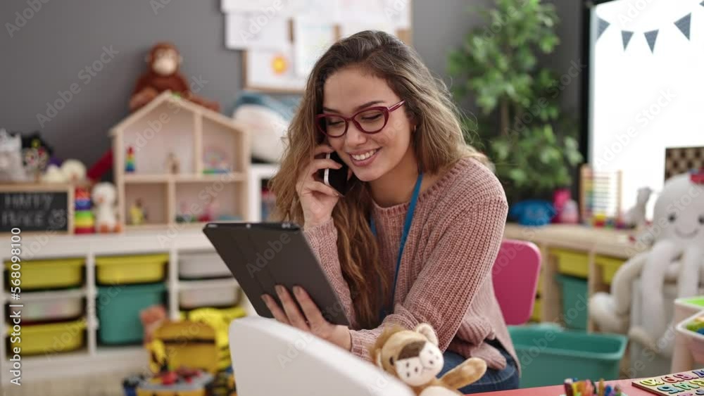Young beautiful hispanic woman preschool teacher talking on smartphone using touchpad at kindergarten Young beautiful hispanic woman preschool teacher talking on smartphone using touchpad at kindergarten