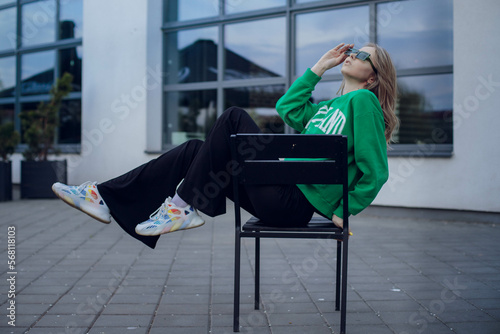 beautiful girl model with coffee in a black jacket in a black baseball cap on a chair sitting outside on the stairs posing for the camera with a phone in green clothes in sneakers fashion