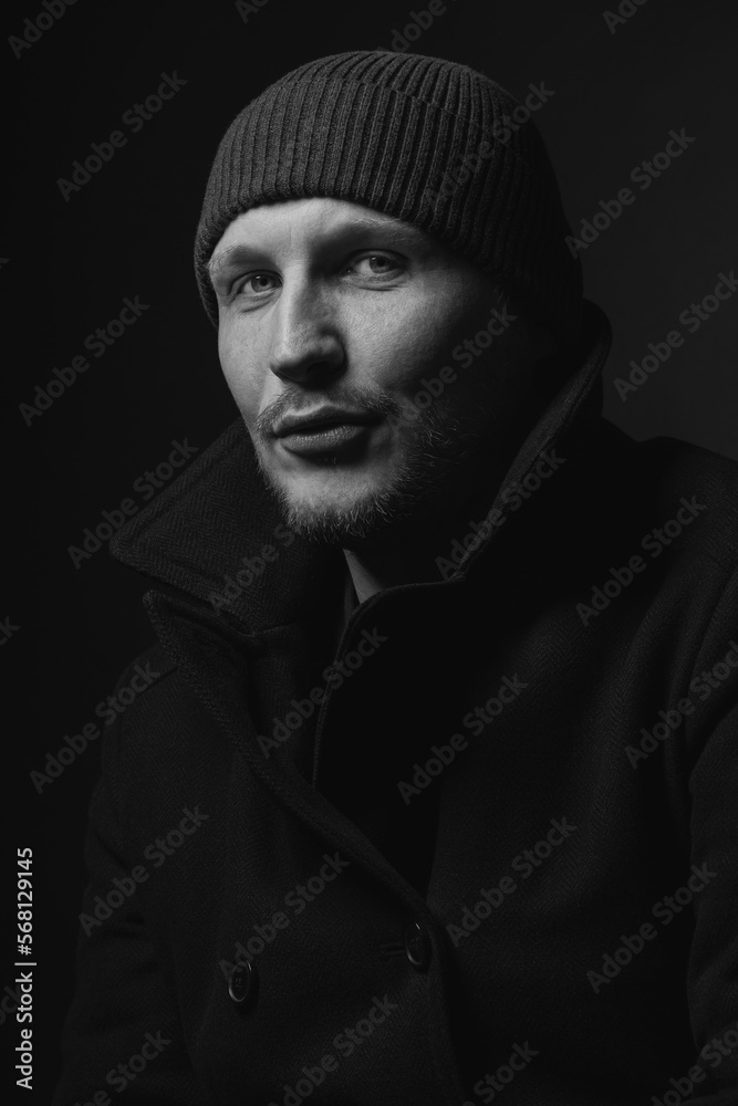 Lifestyle, fashion, occupation concept. Man with winter hat and black coat studio portrait. Model with beard looking at camera with serious look. Dark studio background. Black and white image