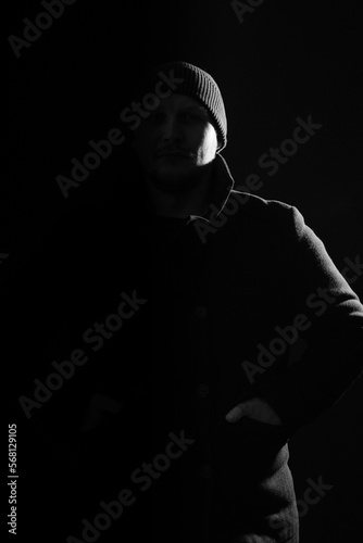 Horror concept. Studio portrait of man with dark coat and hat hiding in black shadow. Model silhouette illuminate with light. Copy space and dark studio background. Black and white image