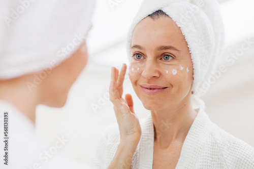 Attractive adult 40s woman apply facial cream middle-aged female look in mirror