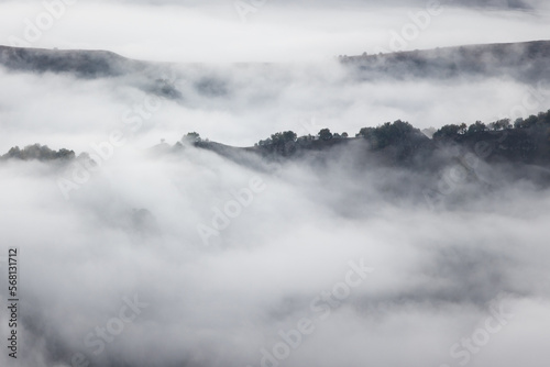Photo of the peaks rock and hills covered trees in early Autumn on the background clouds and fog. Fog lies in the gorges and canyons but peaks see from a fog
