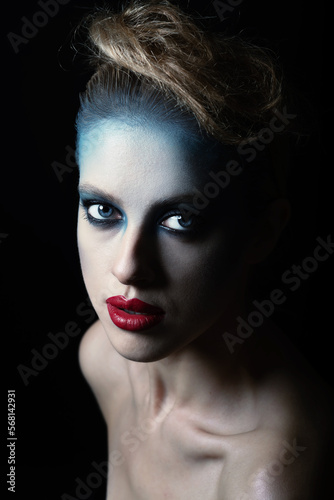 Studio portrait of beautiful woman with fancy and futuristic blue, gray make-up and red lipstick. Model with blue eyes looking at camera with seductive look. Toned image with dark blue color