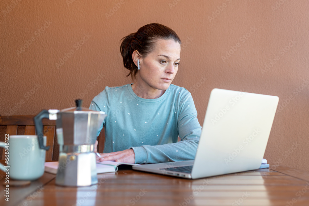 Woman sitting in her pyjamas working on her laptop and writing in a notebook.