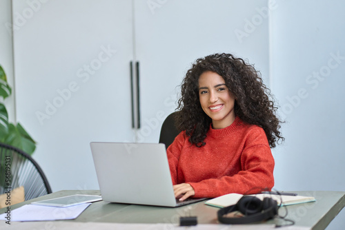 Young happy pretty latin business woman company worker sitting at desk working on laptop. Smiling female professional executive using computer in corporate office looking at camera. Portrait.