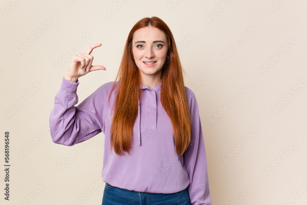 Young red hair woman isolated holding something little with forefingers, smiling and confident.