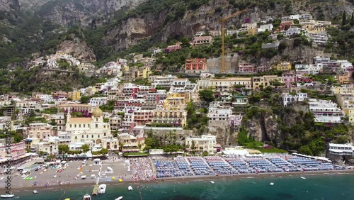 Aerial drone footage of Marina Grande beach, chiesa (church) of Santa Maria Assunta in Positano, Amalfi coast, Campania, Italy.Parasol, sun loungers, people swimming, colourful cliff villas from above