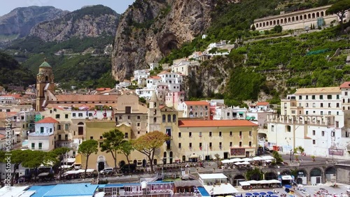 Aerial drone footage of Marina Grande beach, town hall, Sant'Andrea duomo bell tower in Amalfi coast, Campania, Italy. Parasol, sun loungers, people swimming and colourful villas on cliff from above.
