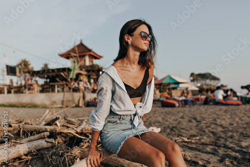 Happy attractive lovely woman with dark short hairstyle wearing denim skirt and light shirt sitting on the ocean shore and enjoying ocean view on sunset 