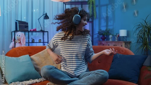 Happy rocker woman with curly hairstyle in wireless headphones relaxing at home dancing on couch listening energetic disco dancing music, playing on imaginary guitar. People weekend leisure activities photo