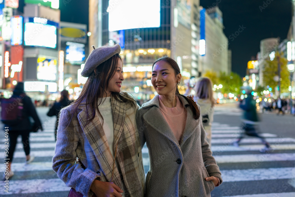 Asian woman shopping and crossing street crosswalk with crowd of people at Shibuya, Tokyo, Japan at night. Happy girl friends enjoy and fun outdoor lifestyle travel in the city in autumn vacation.
