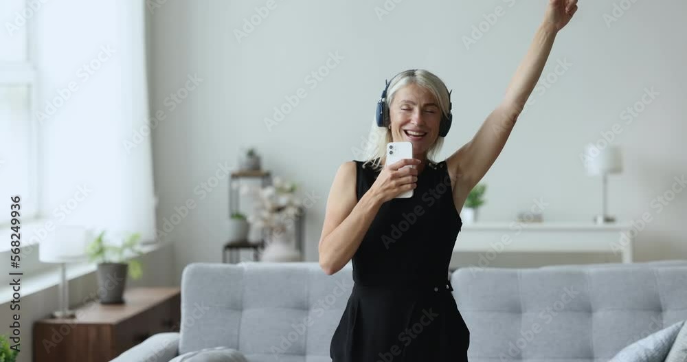 Mature 45s woman wear wireless headphones holds mobile phone singing popular song moving to music standing in living room spend free time, carefree active pastime at home. Karaoke, modern technologies