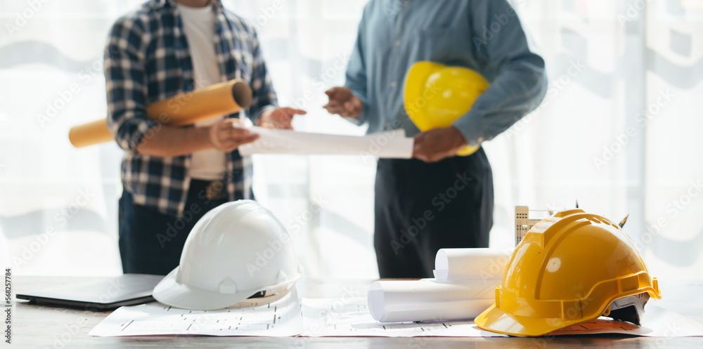 Engineer workspace with safety helmet and blue prints, engineer and teams working in the background.