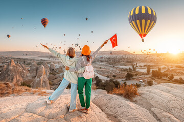 Girl friends travellers with turkish flag, hugging on a viewpoint and admiring view of flying hot air balloons in Cappadocia