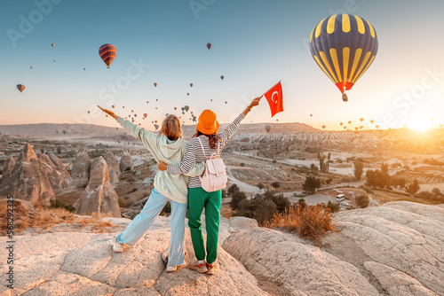 Girl friends travellers with turkish flag, hugging on a viewpoint and admiring view of flying hot air balloons in Cappadocia