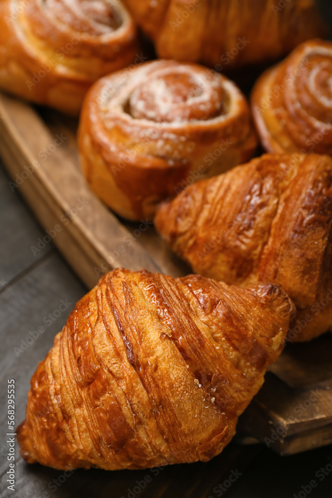 Board with delicious croissants and cinnamon rolls on black wooden background, closeup