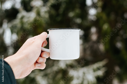 Iron white mug mockup. Close-up of caucasian male hand showing an enamel mug with an empty space on background of pine tree on winter day