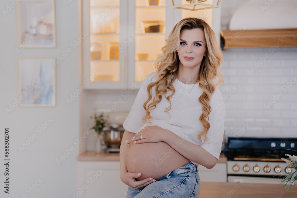 Gorgeous swedish blonde woman with wavy hair sitting on a table at the kitchen looking at camera dreamily. Happy woman waiting for a kid. Maternity and pregnancy concept. Caucasian female at home.