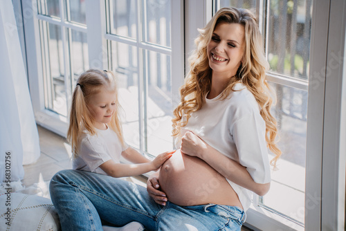 Little cute caucasian girl with ponytails touches pregnant moms belly waits for newborn brother. Cheerful pregnant. Swedish woman sitting on floor at window on sunny day home. Happy pregnancy.