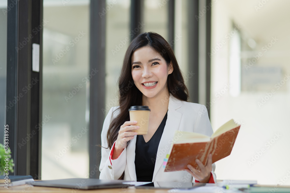 Friendly cheerful asian businesswoman reading a book and holding a coffee mug, people emotions and casual concept, positive woman working in office workspace. Friendly cheerful asian businesswoman reading a book and holding a coffee mug, people emotions and casual concept, positive woman working in office workspace.
