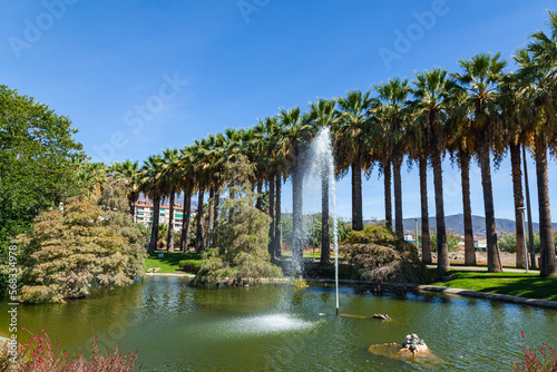 Pueblos de America Park in Motril, Spain, with a fountain spouting water in the middle of a lake and tall palm trees.