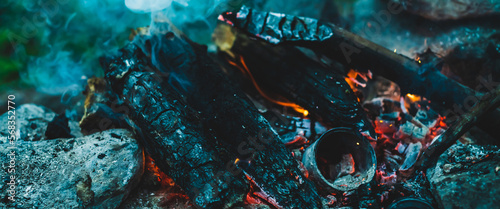 Vivid smoldered firewoods burned in fire close-up. Atmospheric warm background with orange flame of campfire and blue smoke. Wonderful full frame image of bonfire. Burning logs in beautiful fire.