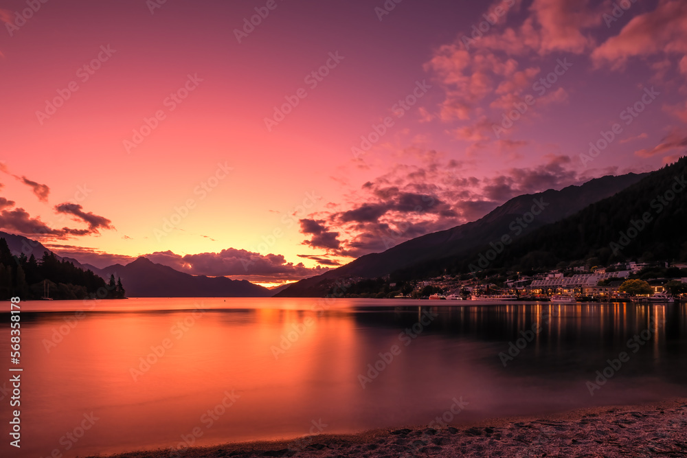 Sunset afterglow lighting up Lake Wakatipu and the town of Queenstown on the South Island of new Zealand with Walter Peak in the distance and beach in the foreground
