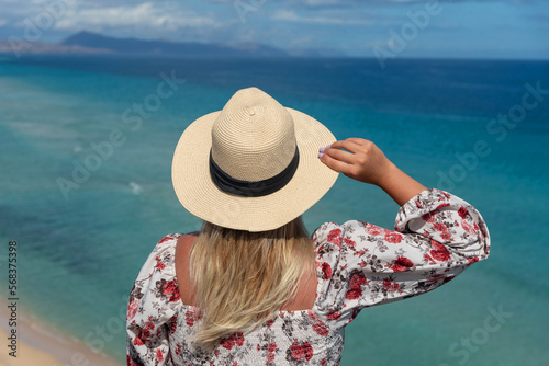 Mujer rubia con el pelo largo en la espalda sosteniendo un sombrero de paja frente al mar turquesa y la playa de arena blanca frente a la costa de Fuerteventura en un día soleado en las Islas canarias