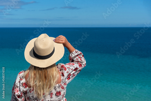 Mujer rubia con el pelo largo en la espalda sosteniendo un sombrero de paja frente al mar turquesa cristalino frente a la costa de la turística Fuerteventura en un día soleado en las Islas Canarias.