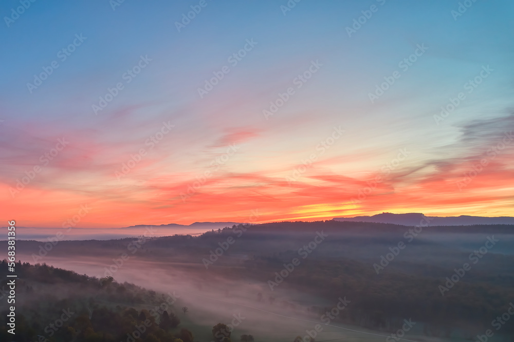 Foggy landscape at sunrise. Range of hills in front of agricultural fields. Blue sky with red accents. Fog in the valley in the autumn season. Aerial view with drone. Germany, Nurtingen, Swabian alb.