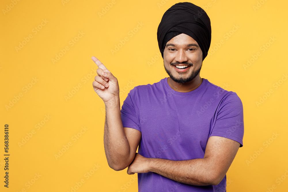 Smiling devotee Sikh Indian man ties his traditional turban dastar wear purple t-shirt point index finger aside on area isolated on plain yellow background studio portrait People lifestyle concept Smiling devotee Sikh Indian man ties his traditional turban dastar wear purple t-shirt point index finger aside on area isolated on plain yellow background studio portrait People lifestyle concept