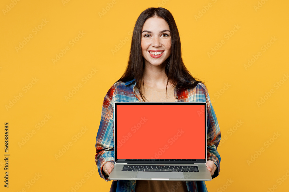 Naklejka premium Young cheerful fun smart smiling IT woman wearing blue shirt beige t-shirt hold use working on laptop pc computer with blank screen workspace area isolated on plain yellow background studio portrait.