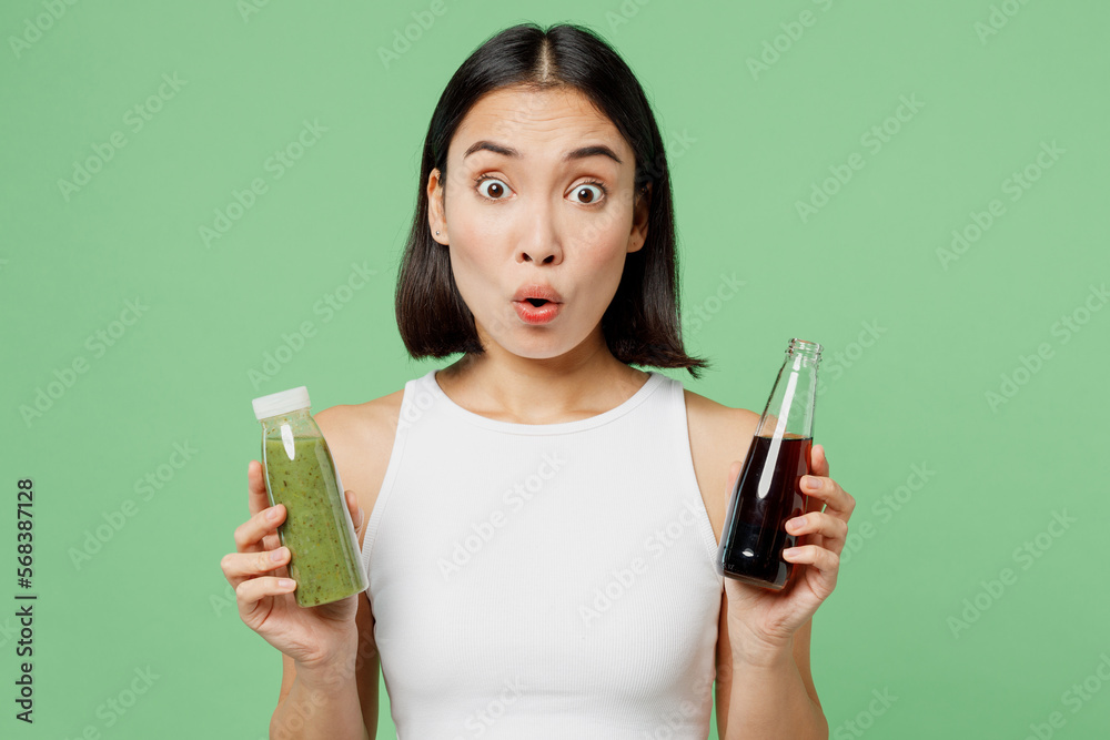 Young shocked woman wearing white clothes hold pressed juice vegetable smoothie soda pop isolated on plain pastel light green background. Proper nutrition healthy fast food unhealthy choice concept. Young shocked woman wearing white clothes hold pressed juice vegetable smoothie soda pop isolated on plain pastel light green background. Proper nutrition healthy fast food unhealthy choice concept.
