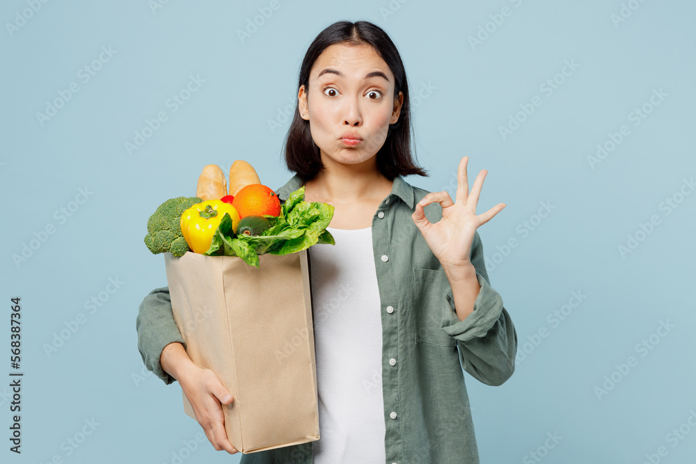 Young satisfied happy woman in casual clothes hold brown paper bag with food products show ok gesture isolated on plain blue cyan background studio portrait. Delivery service from shop or restaurant Young satisfied happy woman in casual clothes hold brown paper bag with food products show ok gesture isolated on plain blue cyan background studio portrait. Delivery service from shop or restaurant