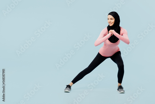 Full body young muslim fitness trainer sporty woman wear pink abaya hijab spend time in home gym do squates lunges look aside isolated on plain blue background studio. Workout sport fit abs concept.
