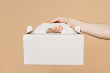 Close up cropped male holding in hand hold cake dessert in unmarked clear empty blank white cardboard box isolated on pastel plain light beige color wall background studio. Service concept concept